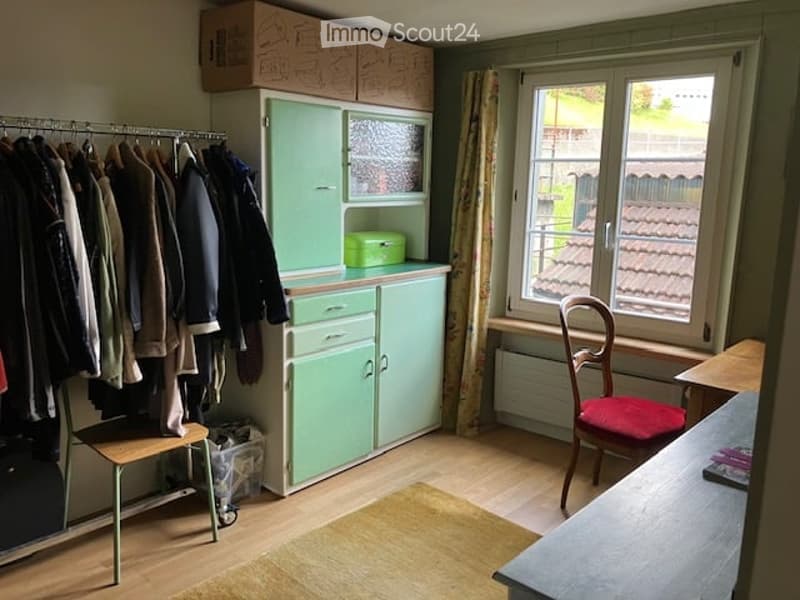 Furnished Room. A small, furnished room with a light green cabinet, a wooden chair with a red cushion, a small table, a coat rack with jackets, and a window with a curtain.