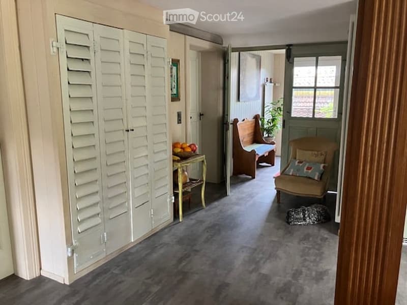 Furnished Room. A well-lit hallway with a grey carpeted floor, featuring a white wardrobe with louvered doors, a small table with fruit, a wooden chair, a sofa, a plant, and a patterned rug.