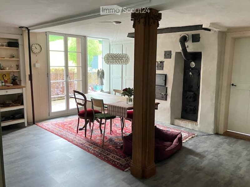 Furnished Room. Bright, open living space with a central dining table and chairs, a large area rug, a dog bed, a shelf with various items, and a clock on the wall. Large glass doors lead to an outdoor area.