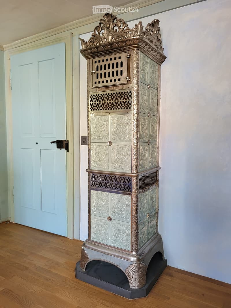 Furnished Room. Ornate, vintage-style metal fireplace with intricate designs and decorative top, placed on a wooden floor in a room with light blue walls and a matching door.