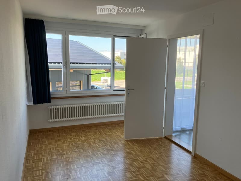Empty room with wooden parquet flooring, white walls, large windows with dark curtains, radiator, and a closed door leading to another space.