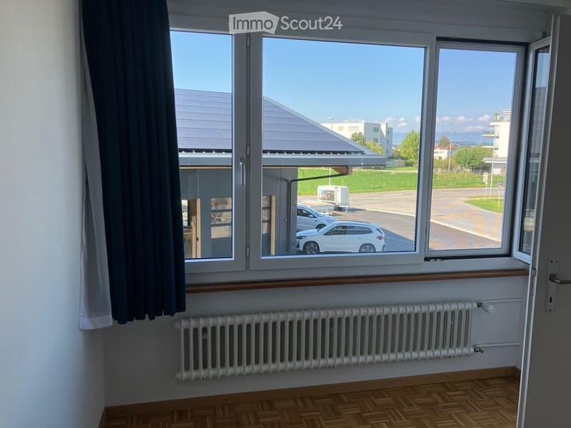 Empty room with wooden flooring, white radiator, large windows with view of parking area and road.