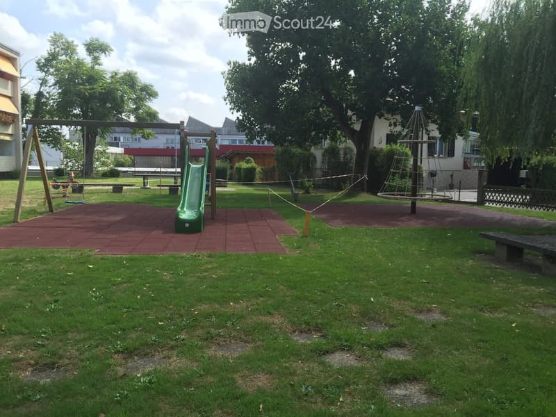 A well-maintained playground area with a green slide, swings, a climbing structure, and benches, surrounded by grass and trees with buildings in the background.