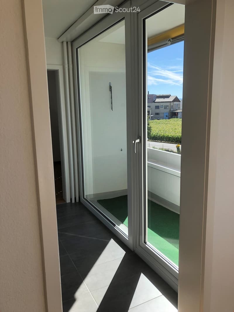 Modern balcony with large glass sliding doors, dark tiled floor, and a view of a green lawn and neighboring buildings.