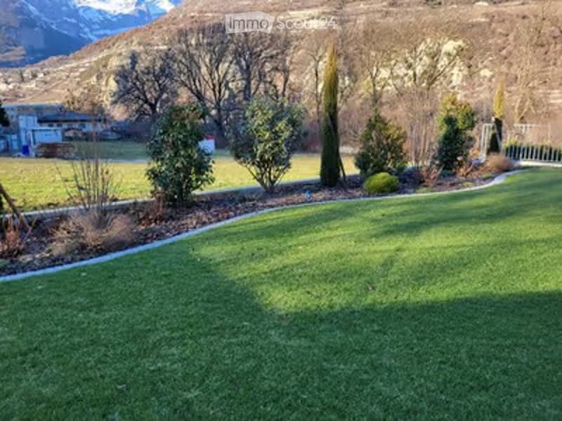 Well-maintained lawn with trimmed edges, surrounded by small shrubs and trees, with a scenic mountain view in the background.