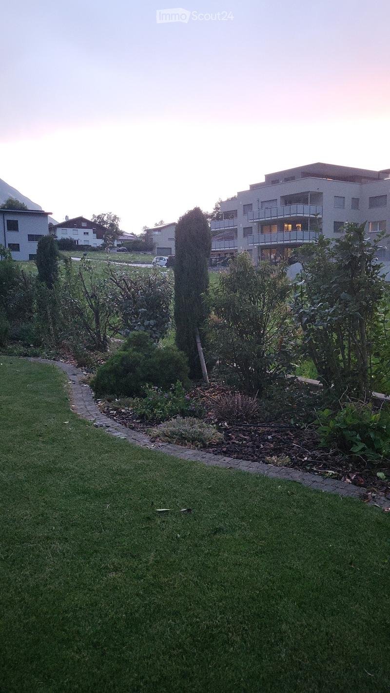 Well-maintained garden with green grass, shrubs, and trees, leading to a modern multi-story building with balconies and large windows.