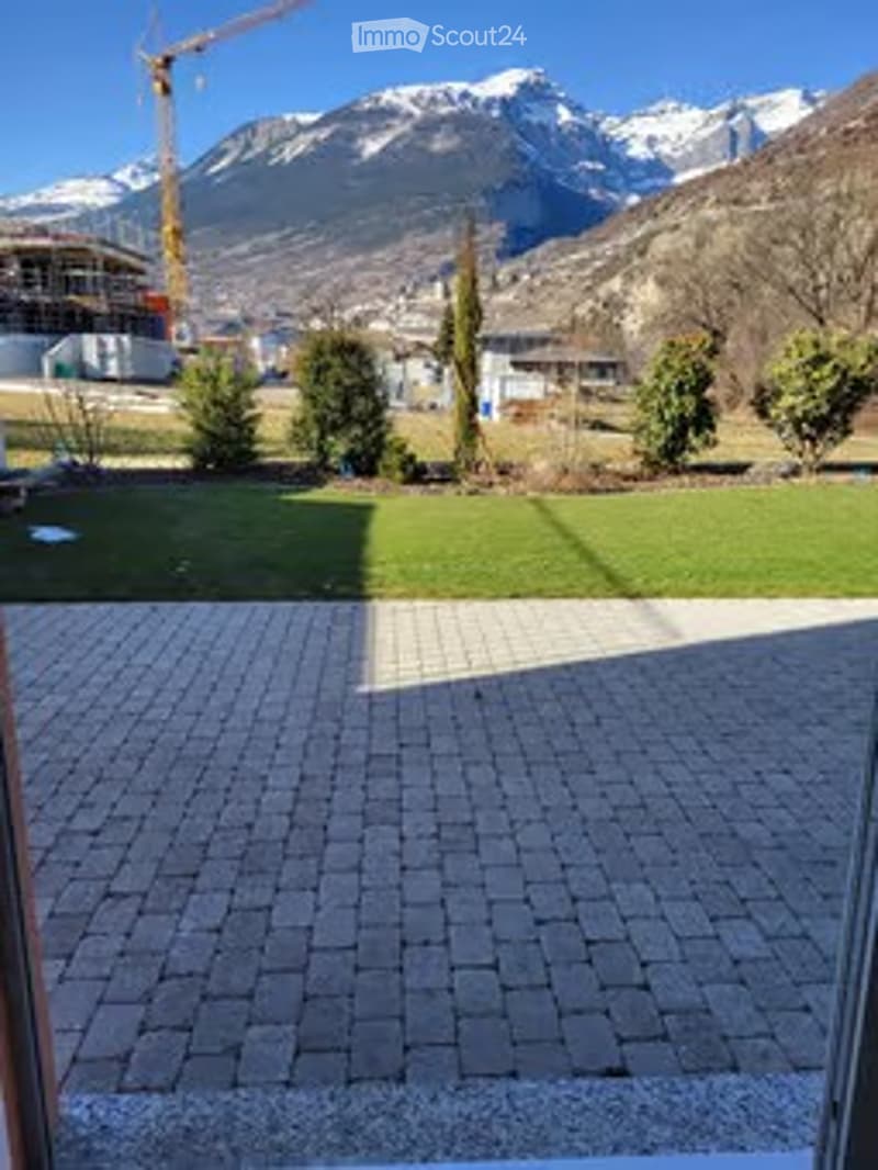 View of a landscaped garden area with a paved brick walkway leading to a grassy lawn, surrounded by trees and distant mountains under a clear blue sky.