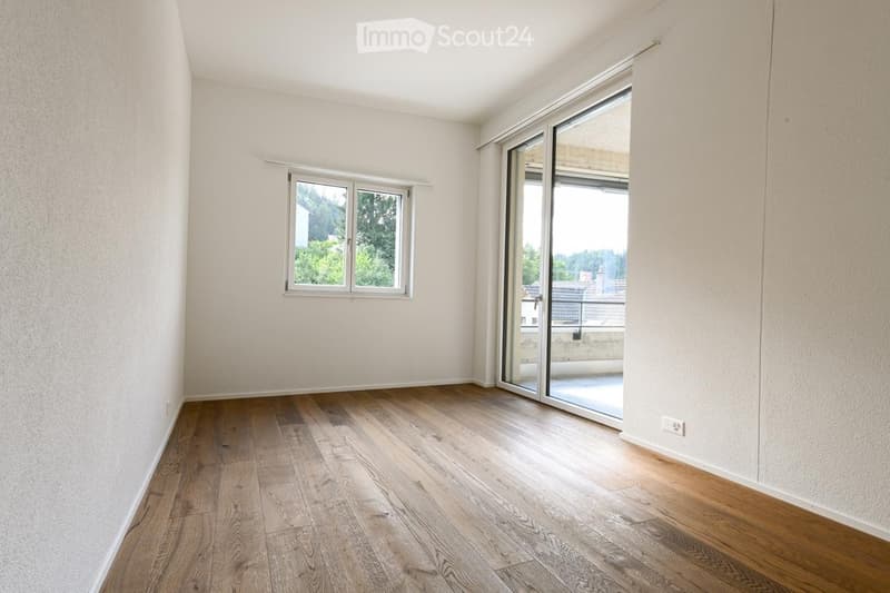 Empty room with wooden flooring, white walls, and large windows including a sliding glass door leading to a balcony.