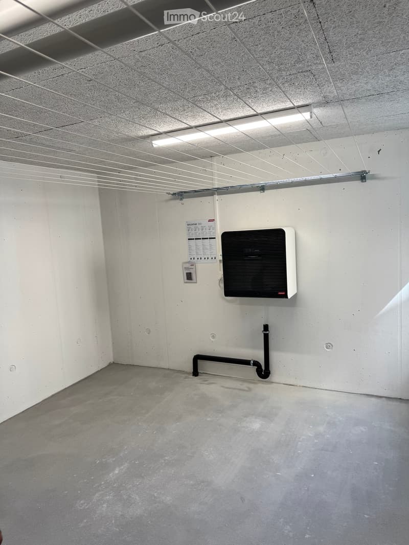 Empty room with white walls, concrete floor, exposed ceiling with lighting fixtures, and a small electrical panel and air conditioning unit mounted on the wall.