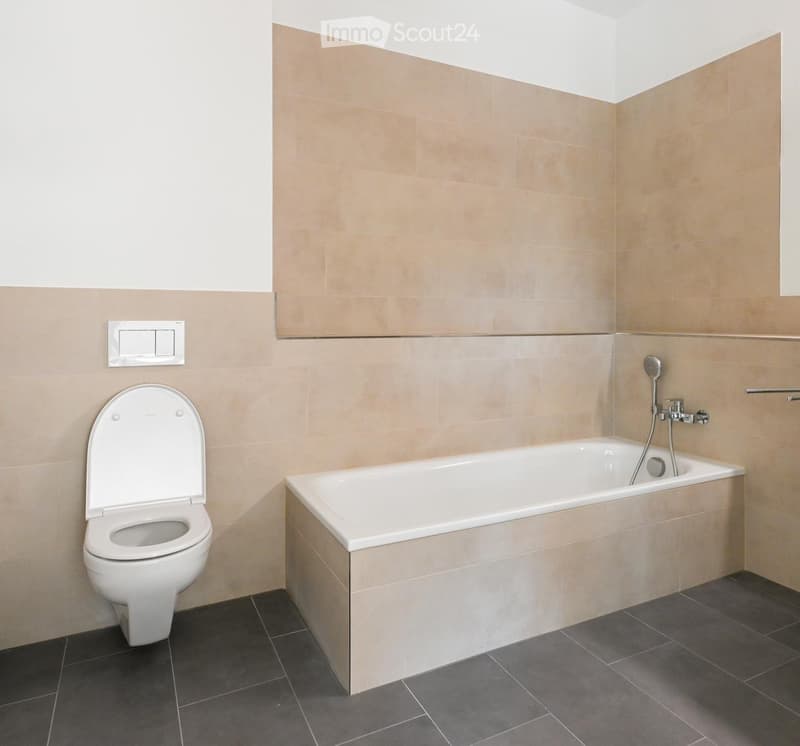 Modern bathroom with a bathtub, toilet, and showerhead, featuring beige tiles and dark grey flooring.