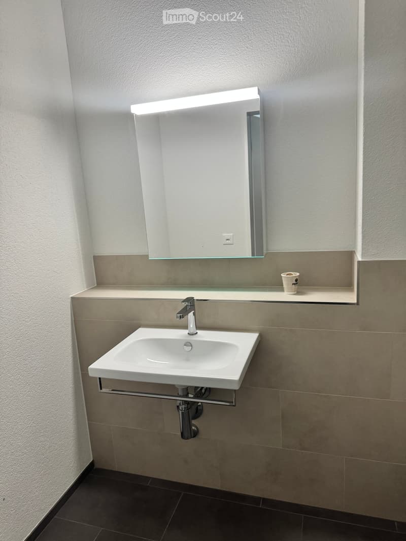 Modern bathroom with a white sink, chrome faucet, illuminated mirror, and a small cup on the counter.