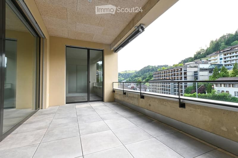 Balcony with tiled floor, glass sliding doors, metal railing, and a view of a cityscape and greenery.