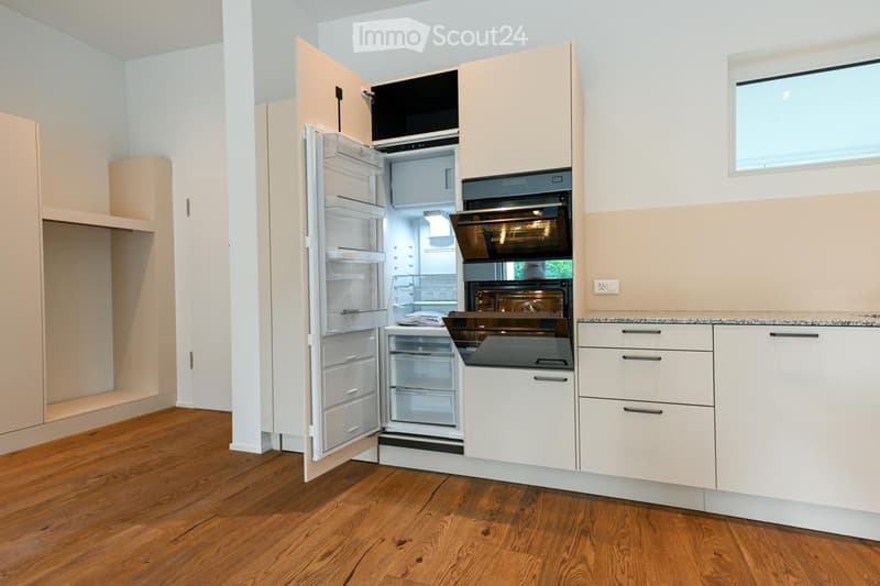 Modern kitchen with light-colored cabinets, a stainless steel refrigerator, and built-in oven. The floor is wooden, and there is a small window above the counter.