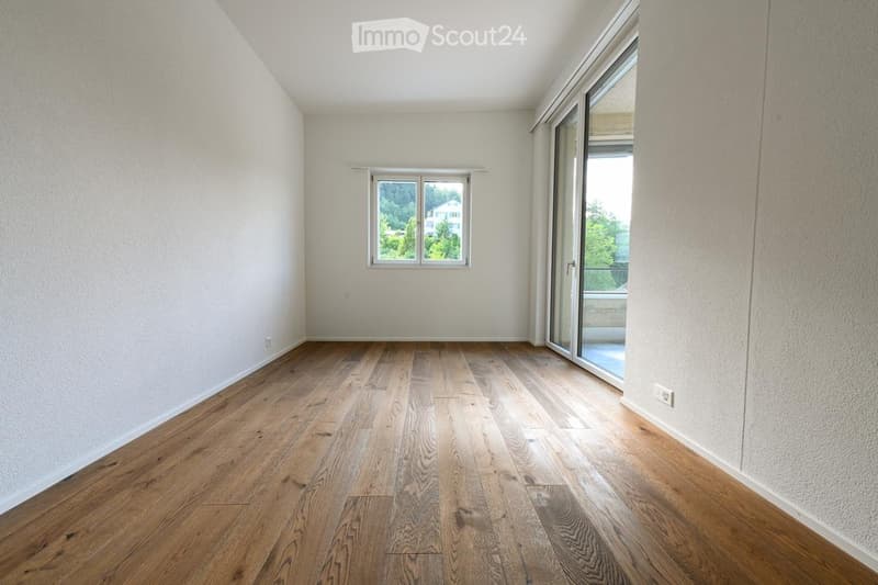Empty room with wooden flooring, white walls, and large windows, including a sliding glass door leading to a balcony.