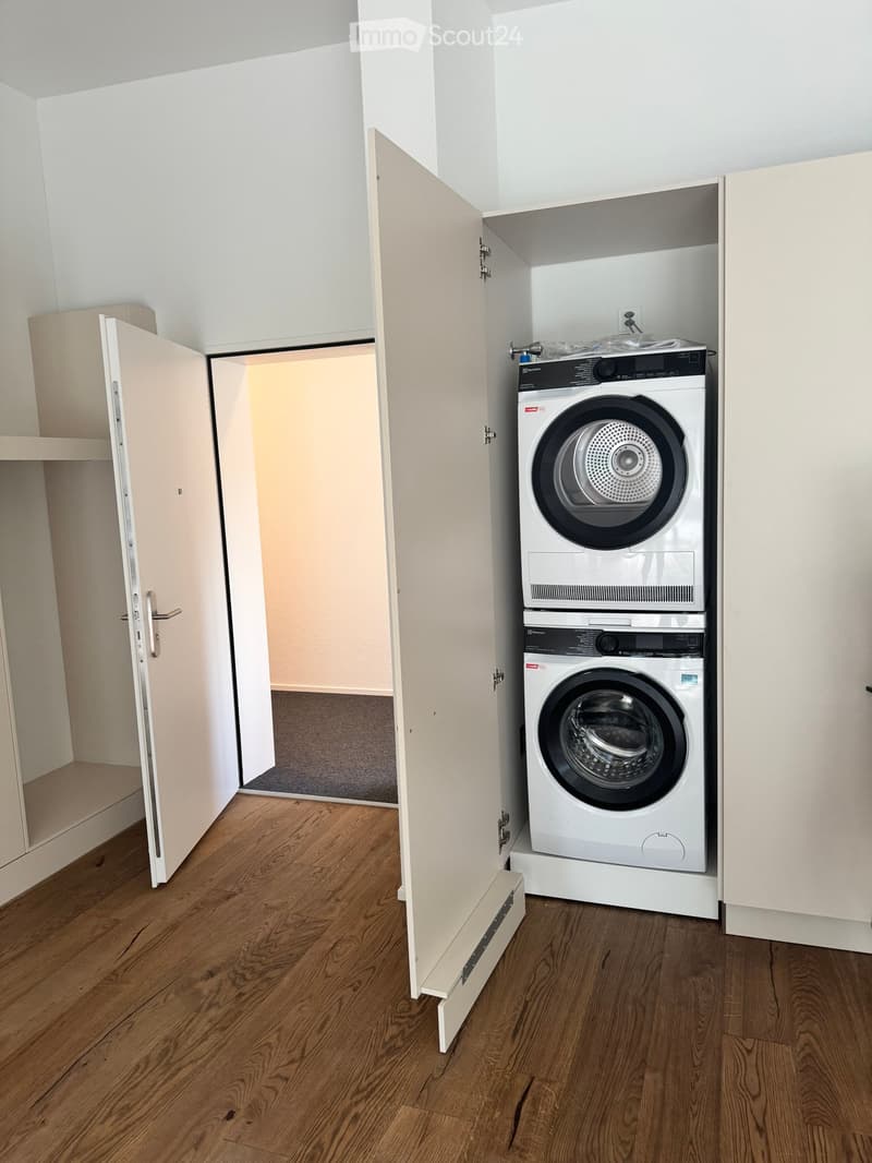 A compact laundry room with a stacked washer and dryer unit, white cabinetry, and wooden flooring.