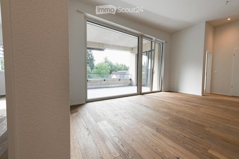 Empty room with wooden flooring, large sliding glass doors leading to a balcony, white walls, and a visible hallway entrance.