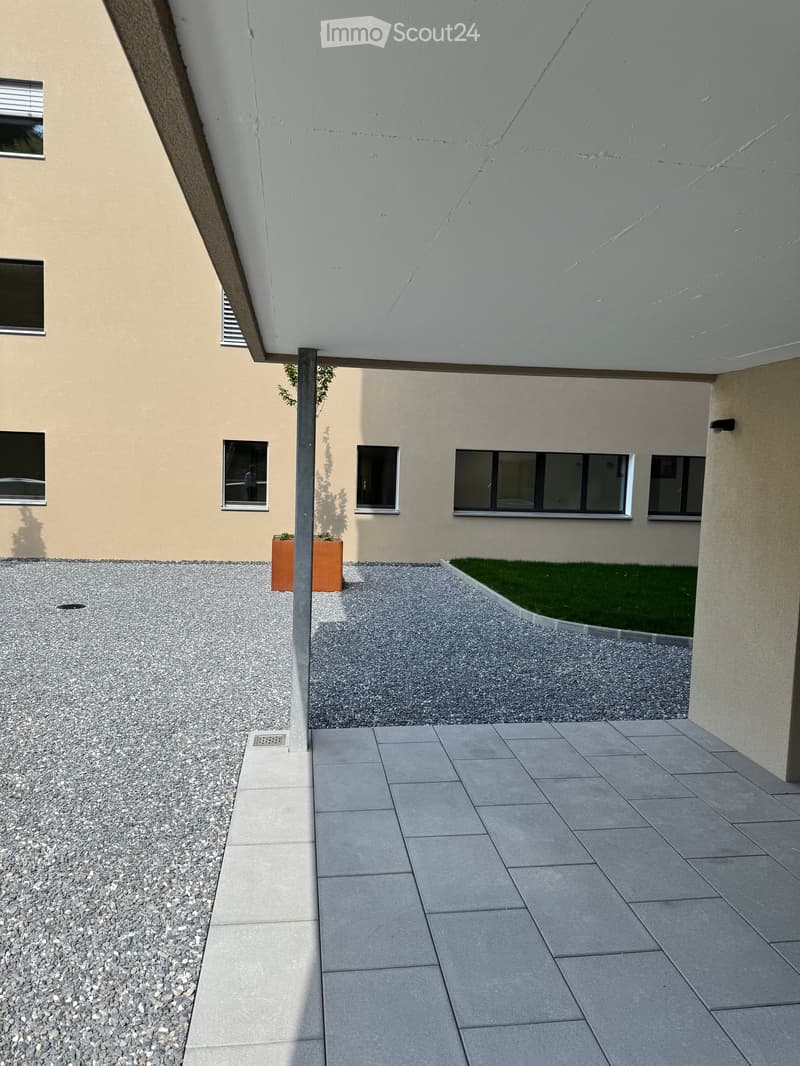 Modern outdoor area with a paved walkway, gravel-covered ground, small garden patch, and potted plants near the building.