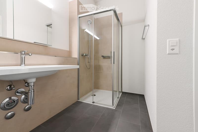Modern bathroom with a white sink, chrome faucet, large mirror, and a glass-enclosed shower with a modern showerhead. The walls are tiled in beige and white, and the floor is dark grey tiles.