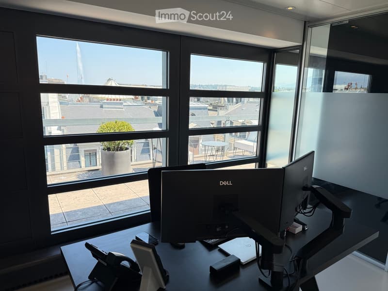 Office space with a large window overlooking a cityscape, featuring a desk with a laptop, monitor, cellphone, telephone, and laptop charger.