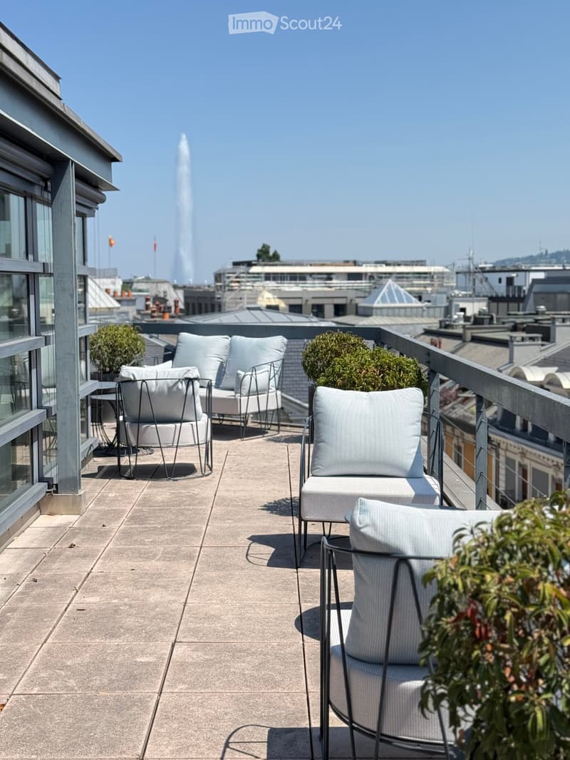 A modern rooftop terrace with light-colored chairs and a small table, overlooking a cityscape with a prominent fountain in the distance.