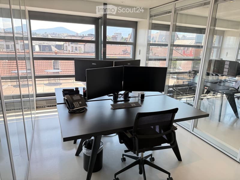 Modern office space with large windows offering a cityscape view, featuring a black desk with multiple computer monitors, a keyboard, a telephone, and an ergonomic office chair.