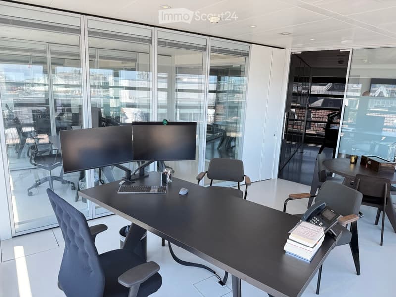 Modern office space with large glass windows, a black desk with dual monitors, ergonomic chairs, and a small round table with a telephone and books.