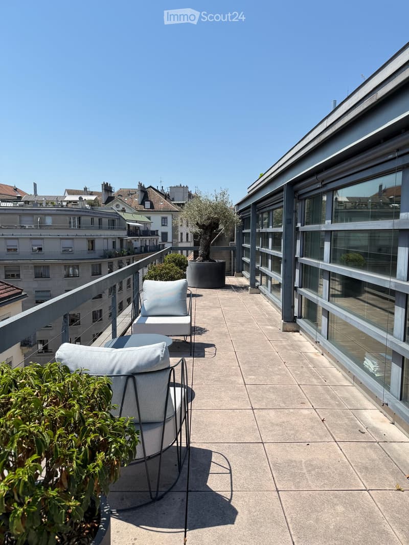 Modern rooftop terrace with a concrete floor, glass railing, and a view of the cityscape. Features a white chair, small table, and potted plants.