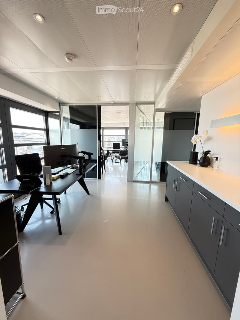 Modern office space with a desk, chair, computer, and cabinet. Large windows provide natural light, and the floor is polished concrete.