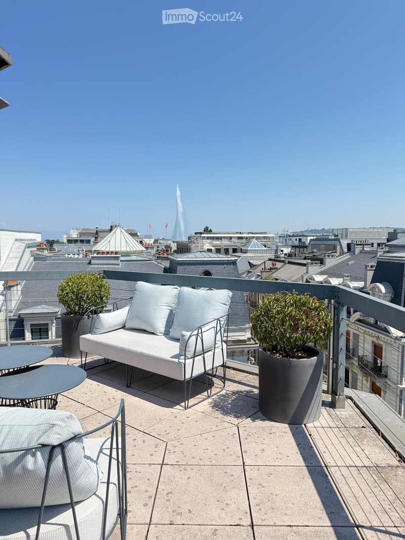 Rooftop terrace with a modern sofa, chair, small table, and potted plants, overlooking a cityscape with visible rooftops and a distant fountain.