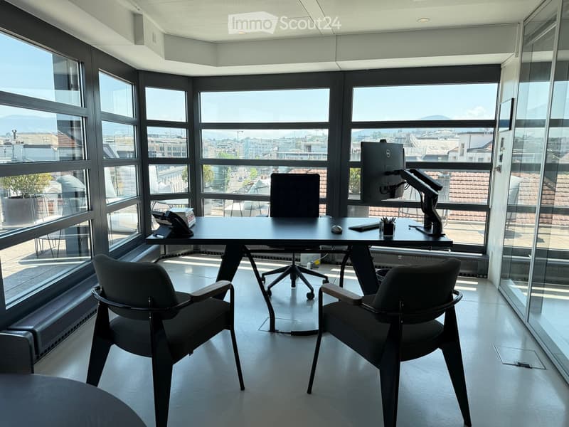 Modern office space with large windows offering a city view, featuring a black desk, two black chairs, a computer monitor, telephone, keyboard, mouse, and pen holder.