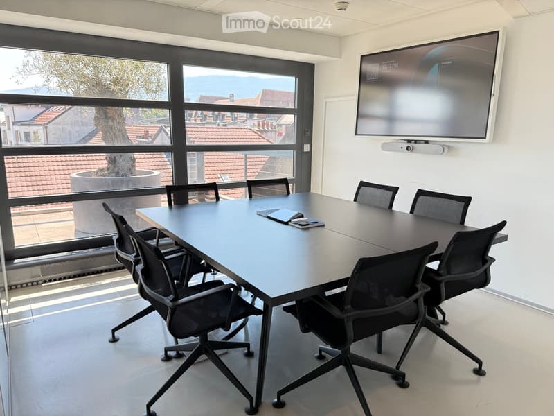 Conference room with a large table, multiple chairs, and a wall-mounted screen. Large windows offer a view of the surrounding area.
