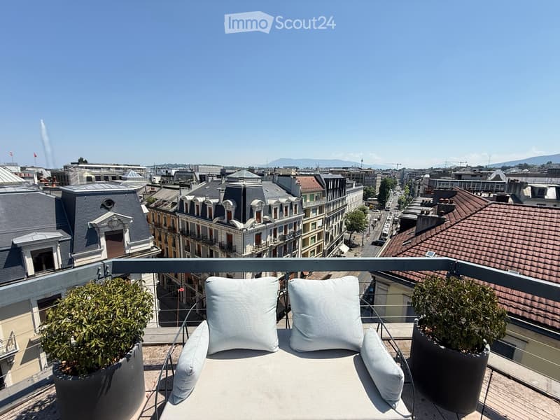 A modern balcony with a comfortable sofa, two white cushions, and two potted plants, overlooking a cityscape with rooftops and distant mountains.