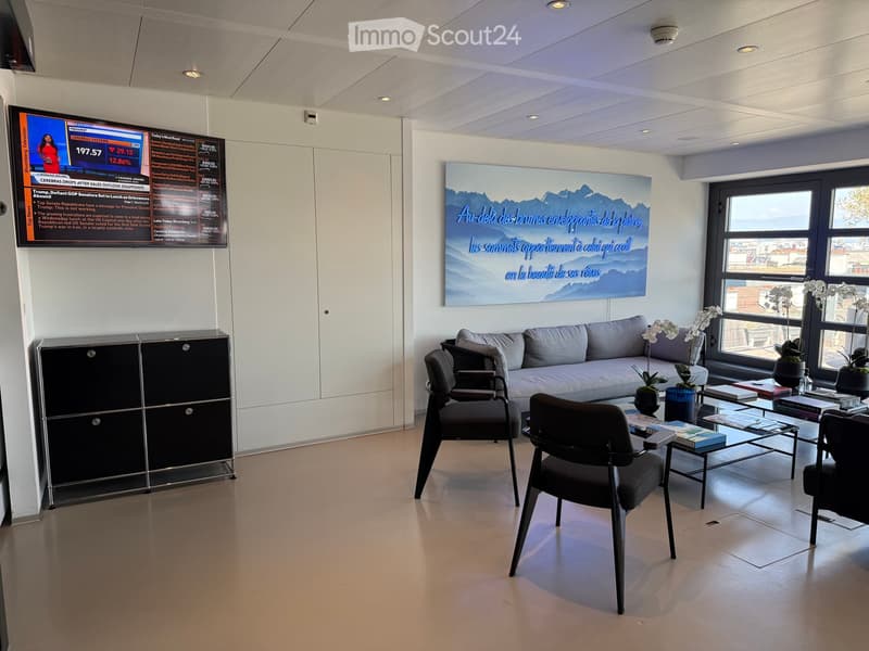 Modern, minimalist living room with a grey sofa, coffee table, chairs, a large screen, and a cabinet. Large windows provide natural light and a view of the city.