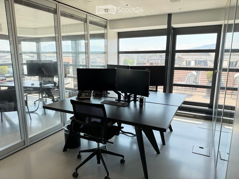 Modern office space with large windows, multiple computer monitors, ergonomic chair, and desk.