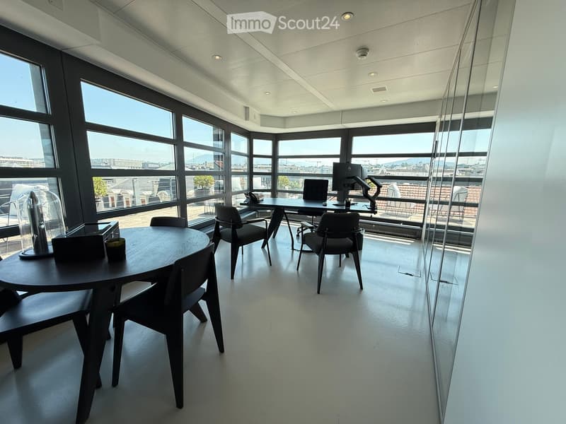 Modern office space with large windows, round table, chairs, computer, and telephone.