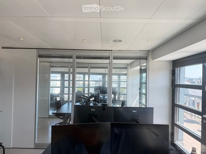 Modern office space with large glass walls, multiple computer monitors, a table, and chairs.