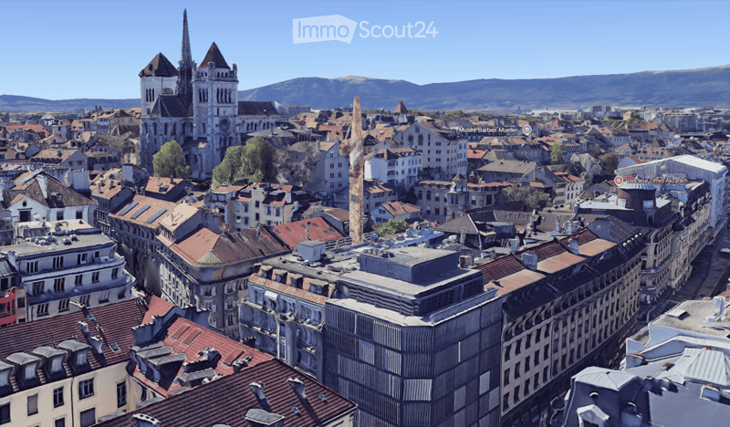 A panoramic view of a historic cityscape with numerous buildings, red-tiled roofs, and a prominent church tower in the background, surrounded by mountains under a clear blue sky.
