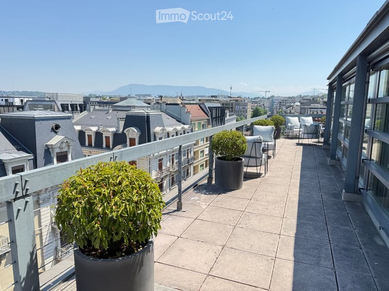 A modern rooftop terrace with a cityscape view, featuring two white chairs, a small table, and two large potted plants.