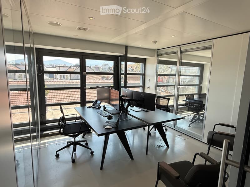 Modern office space with large windows offering a city view, featuring a black desk with two computer monitors, a laptop, a telephone, and several office chairs.