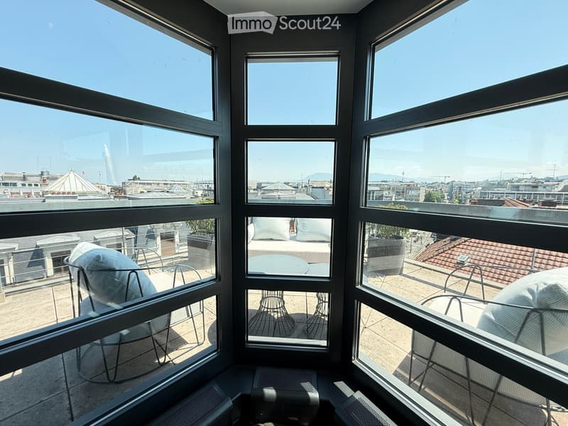 Balcony with large windows overlooking a cityscape, featuring outdoor furniture including chairs and a table.