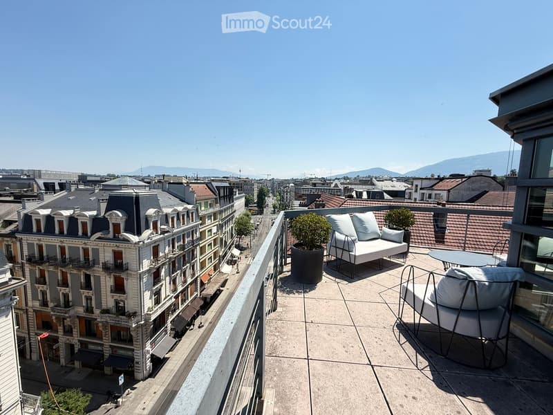 Balcony with a modern sofa, chairs, a small table, and potted plants, overlooking a cityscape with historic buildings and distant mountains.