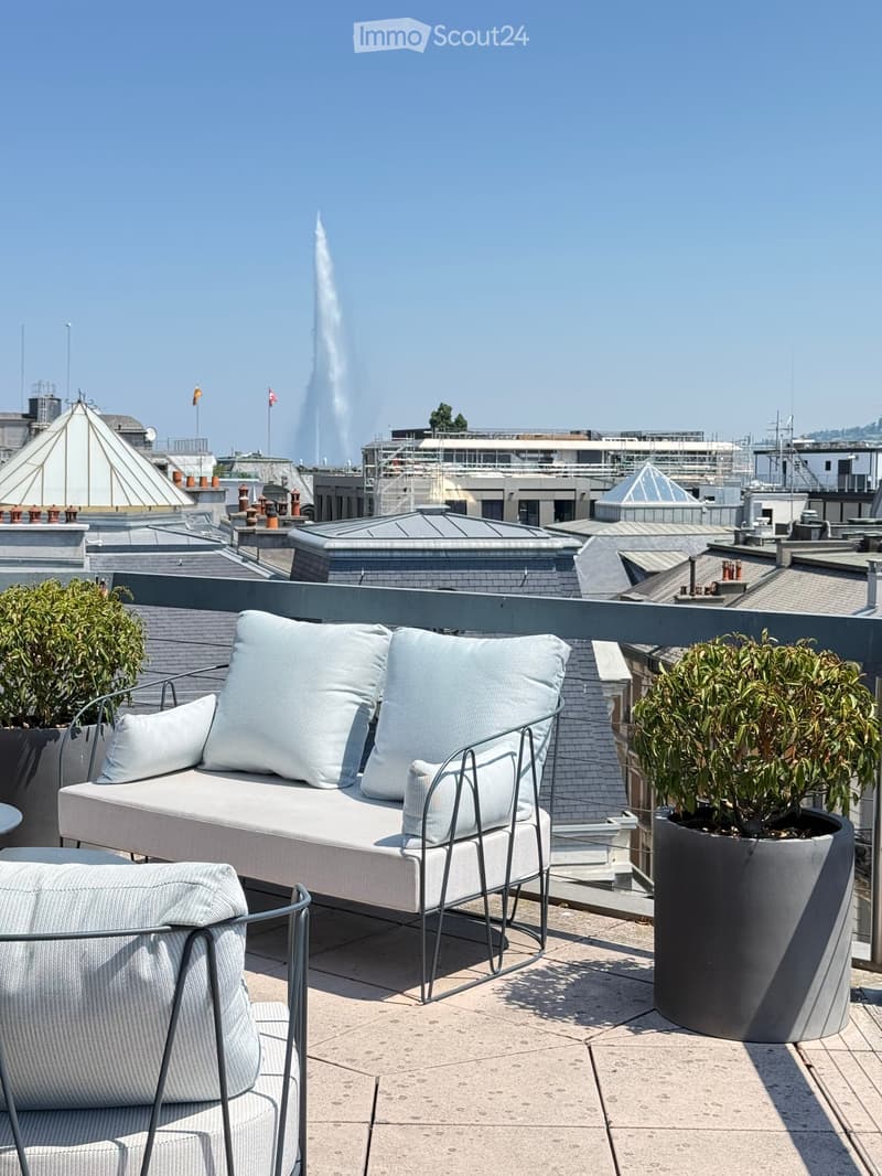 Rooftop terrace with a white sofa, matching chair, and two large potted plants, overlooking a cityscape with a prominent fountain in the background.