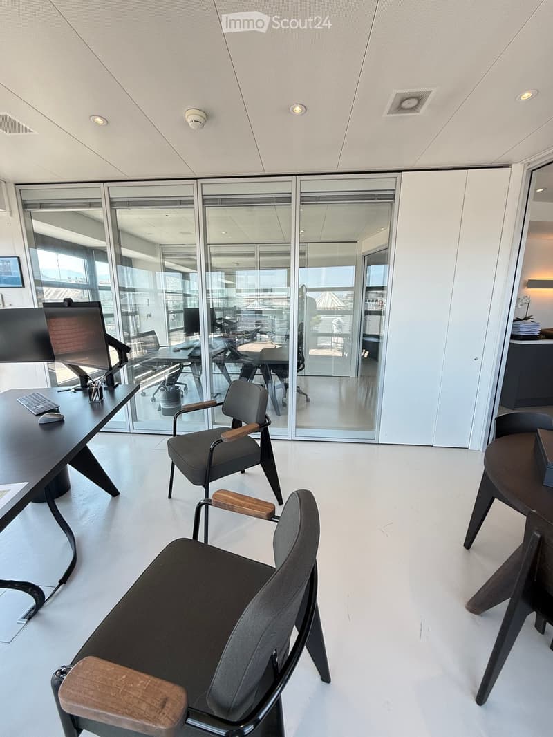 Modern office space with white walls, large glass windows, and contemporary furniture including chairs, tables, and a computer setup.