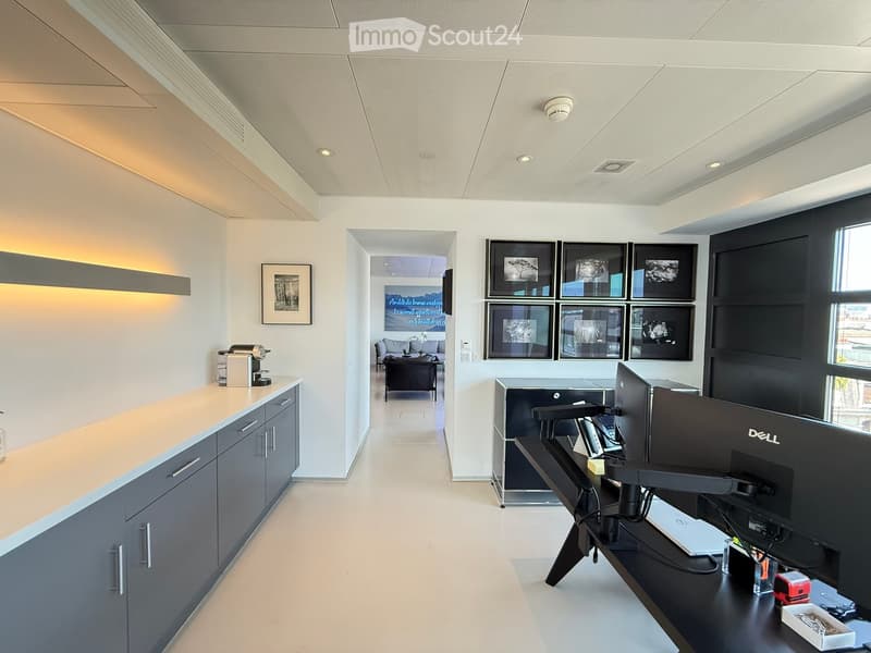 Modern office space with a reception area, desks, chairs, laptops, and a coffee machine. The area features white walls, a sleek reception counter, and framed artwork.