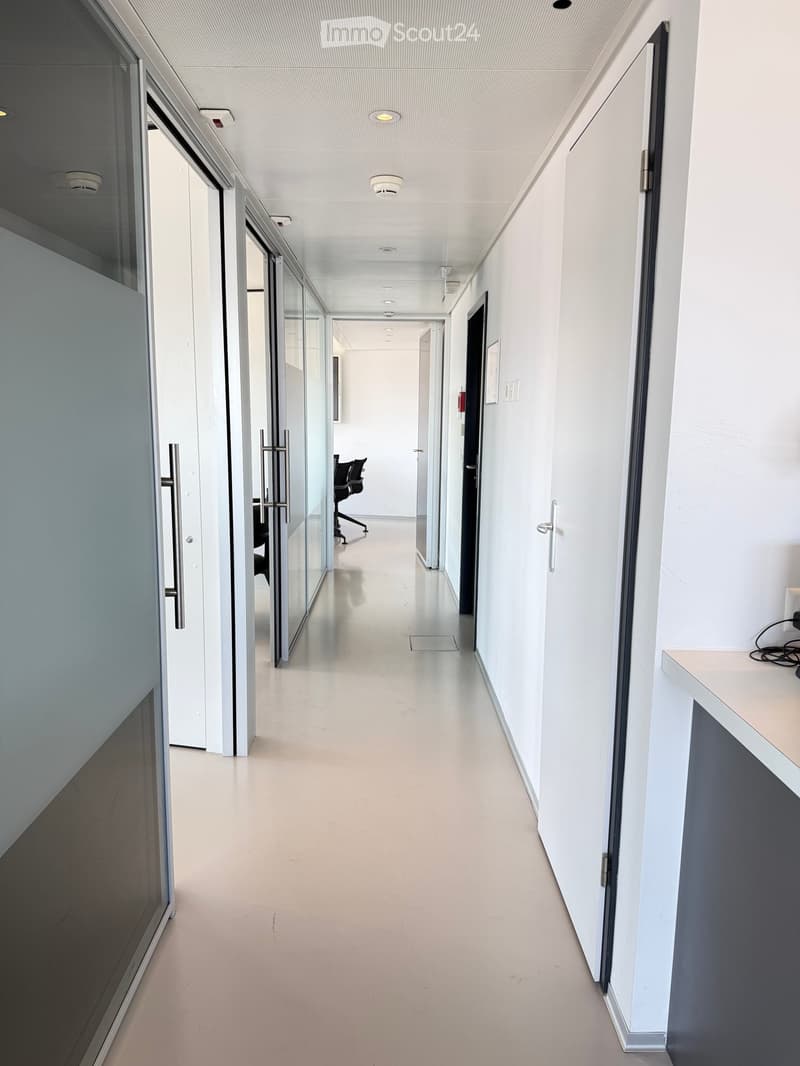 Modern, clean hallway with white walls, light-colored flooring, glass partitions, and recessed lighting. Features a small desk with a computer on the right side.