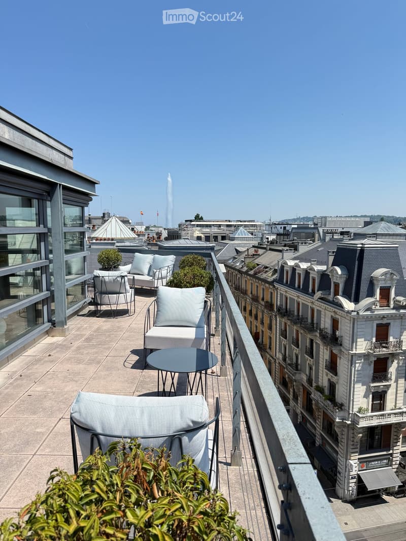 Rooftop terrace with modern furniture, including a sofa, chairs, a small table, and potted plants, overlooking a cityscape with distinctive rooftops and a fountain in the distance.