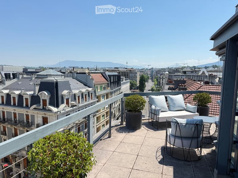 Balcony with a modern seating area, including a sofa, two chairs, a small table, and potted plants, overlooking a cityscape with rooftops and distant mountains.