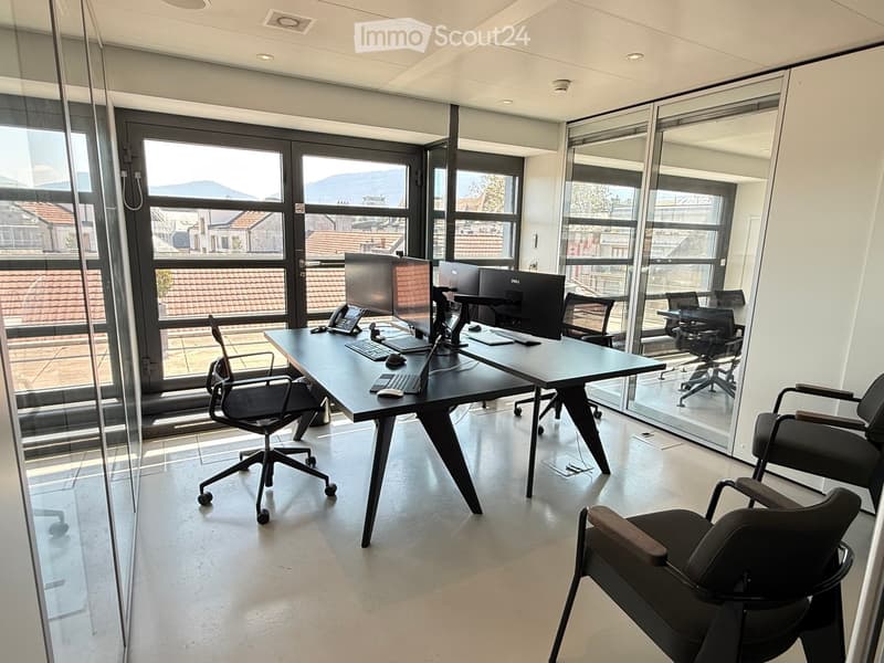 Modern office space with large windows, a black desk, multiple chairs, and computer equipment.