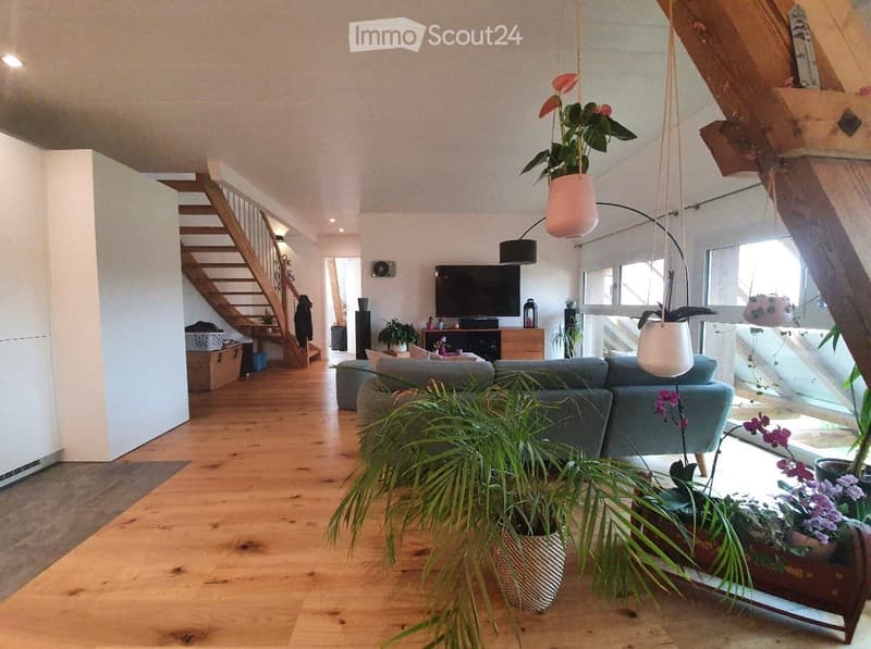 Modern living room with wooden flooring, grey sofa, coffee table, television, floor lamp, and several potted plants. A wooden staircase leads to an upper level.