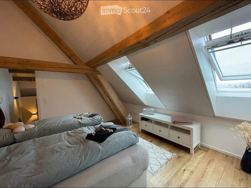 Cozy attic bedroom with wooden beams, two beds, a nightstand, and a small table with a lantern and folded towels.